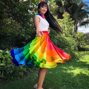 May include: A woman wearing a white sleeveless top and a rainbow-colored skirt with a ruffled hem. She is standing in a grassy area with trees in the background. The skirt is a vibrant rainbow with red, orange, yellow, green, blue, indigo, and violet stripes.