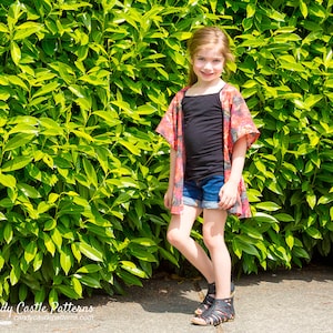 Op de afbeelding: Een jong persoon draagt ​​een kimono met bloemenprint over een zwart tanktop en een spijkerbroek. De persoon staat voor een groene haag.