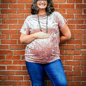 Puede incluir: Una mujer con una blusa de terciopelo rosa con un collar plateado y pantalones vaqueros azules, de pie frente a una pared de ladrillo.