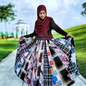 Op de afbeelding: Een kleurrijke patchwork rok met verschillende patronen, waaronder ruitjes en bloemmotieven. De rok wordt omhooggehouden, waardoor de volle, vloeiende vorm zichtbaar is. De persoon draagt een bordeauxrode top en hoofdbedekking.