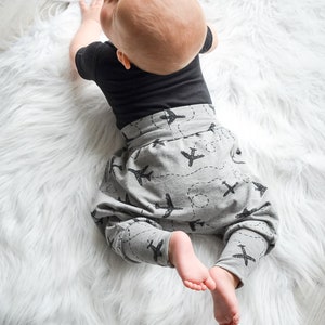 Op de afbeelding: Een baby draagt een grijze broek met vliegtuigprint in zwart en een zwart onesie. De baby ligt op een wit, pluche kleed.