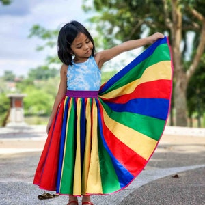 Op de afbeelding: Een jong meisje draagt een regenboog gestreepte rok met een blauwe tanktop en rode schoenen. Ze houdt de rok opzij, waardoor het kleurrijke ontwerp zichtbaar is.