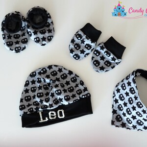 May include: A baby boy's hat, booties, mittens, and bib set in a grey and black skull and star pattern. The hat has the name "Leo" embroidered on the brim.