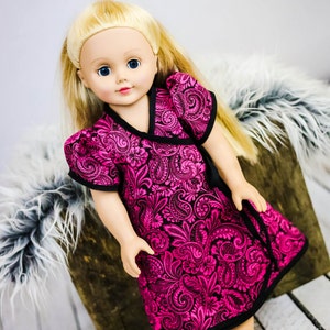 May include: A pink and black patterned dress with a black trim, worn by a doll with blonde hair and blue eyes. The doll is sitting on a wooden surface with a white furry surface behind it. The image includes the text "Candy Castle Patterns".