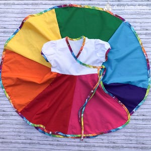Op de afbeelding: Een kleurrijke regenboogjurk met een witte top en een wikkelontwerp. De rok is verdeeld in secties van rood, roze, oranje, geel, groen, blauw en paars. De jurk is afgewerkt met een regenboogpatroon.