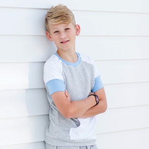 Op de afbeelding: Een jong persoon draagt een grijs en wit shirt met blauwe mouwen en een grijze joggingbroek met een zwart "PEAK" logo. De persoon staat tegen een witte muur.