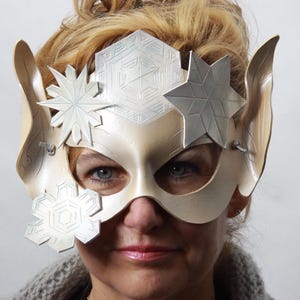 May include: A cream-colored masquerade mask featuring large pointed elf-like ears on the sides. The mask is decorated with silver-toned geometric snowflake and star-shaped embellishments across the forehead and near the eye openings.