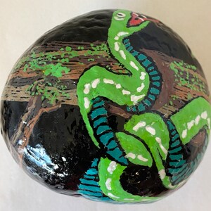 May include: Hand-painted rock with a green snake design. The snake is curled up on a black background with brown and green accents.