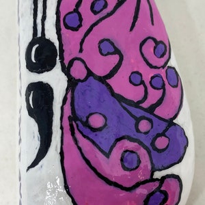 Può includere: Sasso dipinto a mano con un disegno a farfalla. La farfalla ha ali viola e rosa con contorni neri. La pietra è bianca con finitura lucida. Un disegno nero è sul lato sinistro.