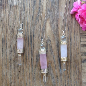 May include: Three silver necklaces with pink crystal pendants. Each pendant is a cylinder with a clear crystal ball at the top and a clear crystal point at the bottom. The necklaces are hanging against a wooden background.
