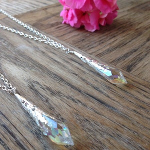 May include: Two silver necklaces with iridescent crystal teardrop pendants hanging on a wooden surface. The necklaces are layered on top of each other.