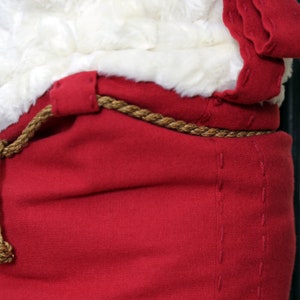 May include: Close-up of a red Santa suit with a white fur trim and a brown rope belt. The suit has a stitched seam detail.