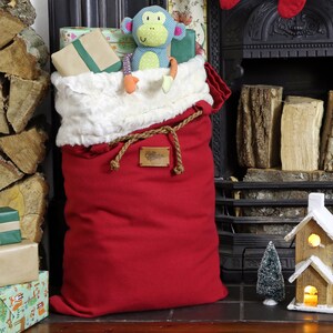 May include: A red Santa sack with a white faux fur trim and a brown rope drawstring. The sack is filled with wrapped gifts and a stuffed monkey toy. The sack is sitting in front of a fireplace.