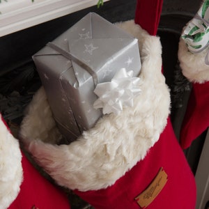 Può includere: Un calzino di Natale rosso con un bordo in pelliccia sintetica bianca è pieno di una scatola regalo argentata avvolta con un fiocco bianco. Il calzino ha un'etichetta in pelle marrone con il testo "Buon Natale" su di essa.