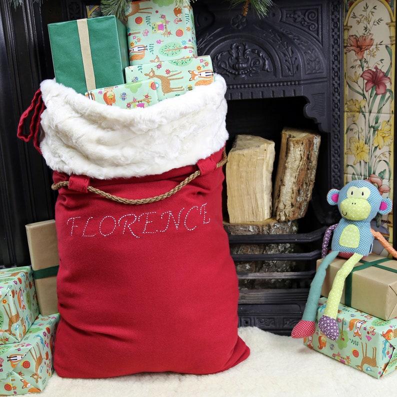 May include: A red Santa sack with a white faux fur trim and the name "FLORENCE" embroidered in white on the front. The sack is filled with wrapped gifts and is sitting in front of a fireplace.