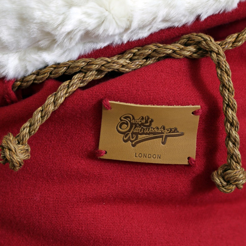 May include: A red fabric bag with a brown rope handle and a brown leather tag that says "Santa's Little Workshop, London".