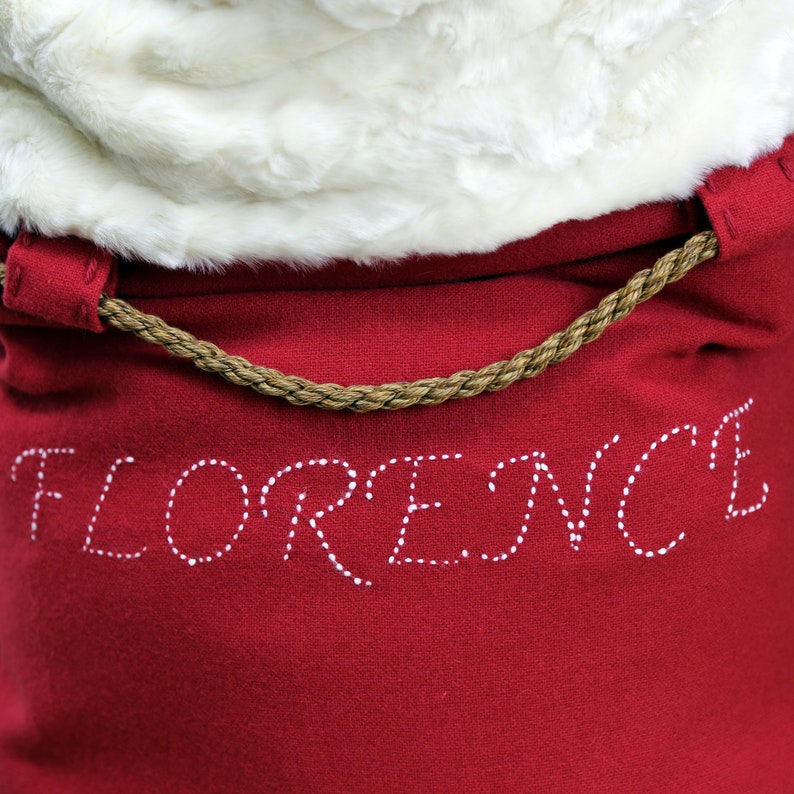 May include: A red fabric bag with a white fur trim and a brown rope handle. The bag has the word "FLORENCE" stitched in white on the front.