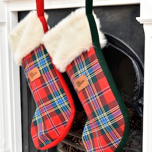 Puede incluir: Dos calcetines de Navidad de cuadros rojos y verdes con ribete de piel sintética blanca y una etiqueta de cuero marrón que dice "Feliz Navidad".