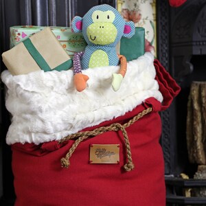 May include: A red Santa sack with a white faux fur trim and a brown rope tie. The sack is filled with gifts and a blue and green monkey toy is peeking out of the top.