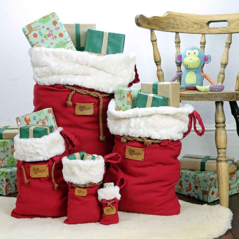 May include: Five red fabric gift bags with white faux fur trim and rope handles. The bags are filled with wrapped gifts and are arranged in a pyramid shape. The largest bag is at the bottom and the smallest bag is at the top. The bags are sitting on a white rug in front of a wooden chair.