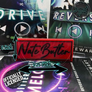 May include: A red rectangular sign with the name "Nate Butter" in black script, measuring 5.25 inches long and 2 inches high. The sign is positioned in front of book covers. The words "Officially Licensed" are also visible.