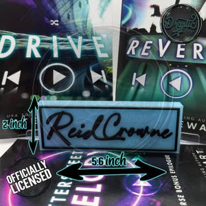May include: A light blue rectangular sign with the name "Reid Crowne" in black script. The sign is 5.6 inches long and 2 inches tall. The background features book covers with the words "DRIVE" and "REVERSE". The sign is officially licensed.