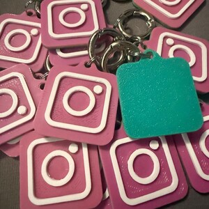 Pode incluir: Uma coleção de chaveiros rosa e brancos com o logotipo do Instagram, com argolas prateadas. Um chaveiro turquesa também está presente. Os chaveiros são quadrados, com uma borda branca em relevo e um ícone de câmera branco no centro.