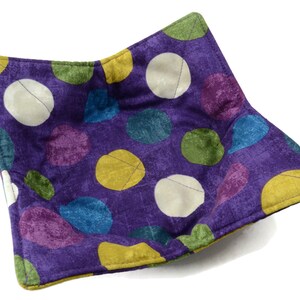 May include: Purple fabric bowl with a colorful geometric pattern of circles and ovals. The bowl is made of a soft, quilted fabric and is perfect for holding small items.