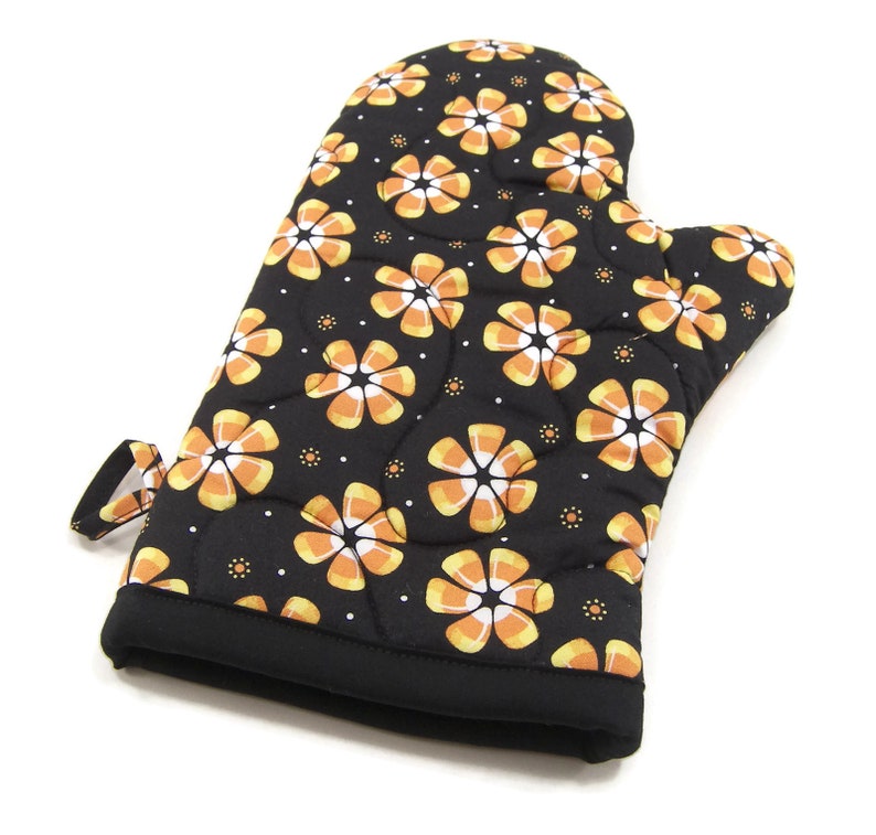 May include: Black oven mitt with a pattern of orange and white candy corn flowers. The mitt has a quilted texture and a hanging loop.