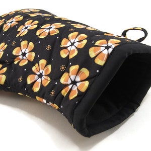 May include: Black oven mitt with a quilted design featuring orange and white candy corn flowers. The mitt has a loop for hanging.