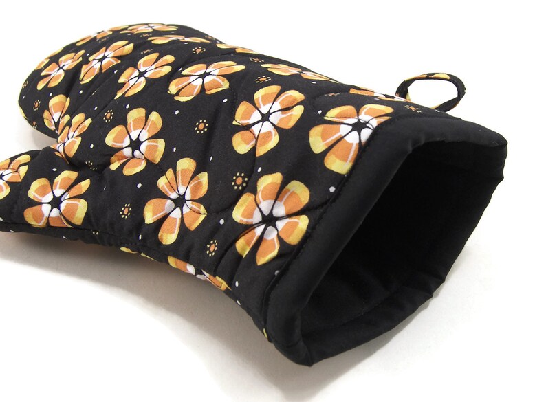 May include: Black oven mitt with a quilted design featuring orange and white candy corn flowers. The mitt has a loop for hanging.