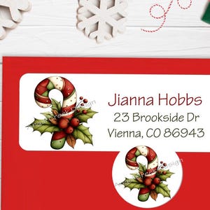 May include: A red Christmas-themed address label with a candy cane and holly design. The label includes the name "Jianna Hobbs" and an address. A matching round sticker with the same design is also present.