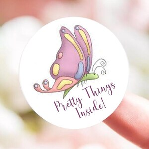 May include: A round white sticker featuring a cartoon butterfly with pink and yellow wings, blue accents, and a green body. The sticker has the text "Pretty Things Inside!" in a purple script font.