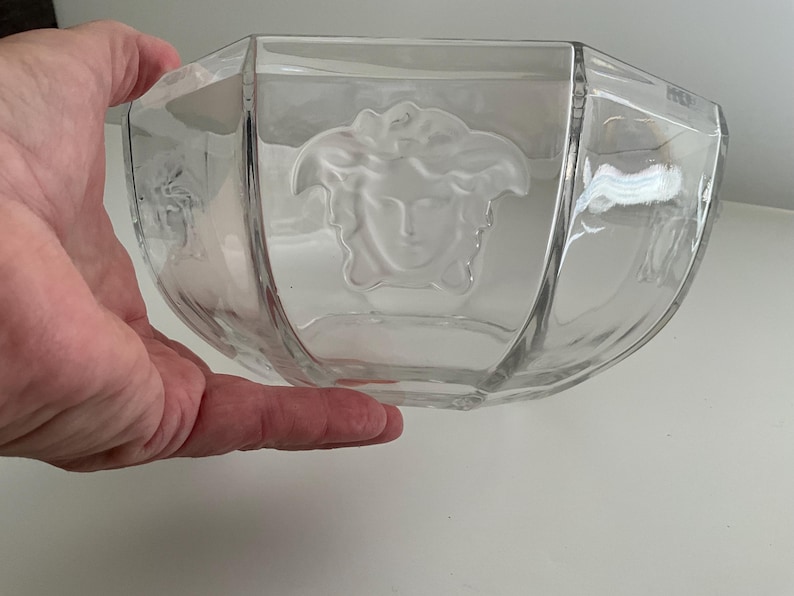 Puede incluir: Cuenco de vidrio transparente con un dise&ntilde;o geom&eacute;trico. El cuenco presenta un emblema de cabeza de Medusa en el centro. El cuenco est&aacute; sostenido por una mano.