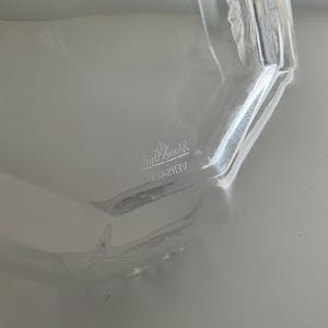 Puede incluir: Contenedor de vidrio transparente con forma geom&eacute;trica. La palabra "VERSACE" es visible en la imagen. El contenedor es transparente y refleja la luz, lo que sugiere una superficie pulida. El dise&ntilde;o es simple y elegante.