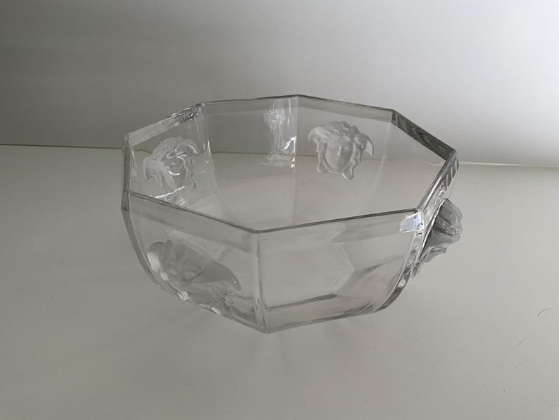 Puede incluir: Cuenco de vidrio transparente, octogonal, con detalles en relieve de cabeza de Medusa. El cuenco tiene un dise&ntilde;o geom&eacute;trico y probablemente se utiliza con fines decorativos o de servicio. El vidrio transparente permite ver el contenido.