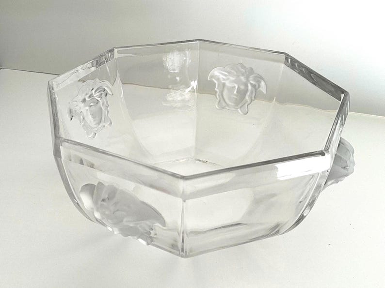 Puede incluir: Cuenco de vidrio transparente, octogonal, con detalles en relieve de cabezas de Medusa. El cuenco tiene un dise&ntilde;o geom&eacute;trico y probablemente se utiliza para servir o exhibir. El vidrio transparente permite la visibilidad del contenido.