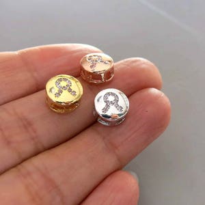 May include: Three round metal charms with a small ribbon design and clear rhinestones. The charms are gold, rose gold, and silver.
