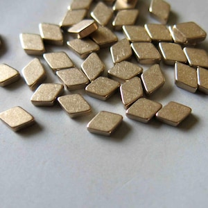 May include: A scattering of small, diamond-shaped metal beads with a matte gold finish. The beads are spread across a light-coloured surface, highlighting their geometric form and metallic sheen. Suitable for jewellery making or craft projects.