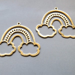 Puede incluir: Dos dijes de metal de color dorado en forma de arcoíris y nubes. Cada dije presenta un arcoíris con un contorno punteado y dos nubes a cada lado.