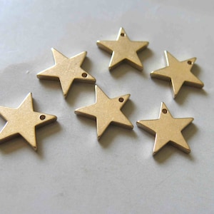 May include: Six gold-colored metal star charms with a hole in the center of each star. The stars are arranged in a semi-circle on a white surface.