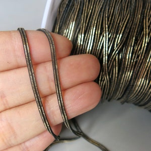 May include: Close-up of a spool of black and gold metallic cord. Two strands of the cord are held, showing the flat, ribbed texture. The cord is likely used for jewellery making or crafting.