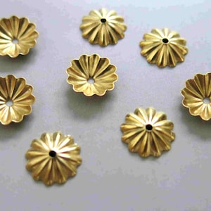 May include: A collection of gold-coloured, flower-shaped metal bead caps. Each cap features a central hole and radiating petal-like ridges. The caps are arranged on a light grey surface.