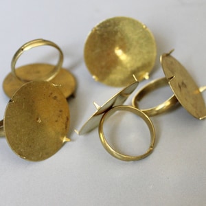 May include: Five gold-toned metal ring bases for setting cabochons or other jewelry components. The bases have a round shape with a raised center and three prongs on the bottom.