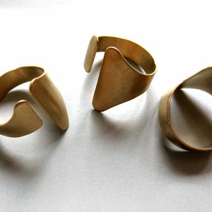 Puede incluir: Tres anillos de metal dorado con diseños únicos. Los anillos son abiertos y tienen un estilo simple y minimalista.