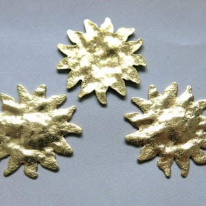 May include: Three gold-coloured, sun-shaped metal embellishments with a textured surface. The sunburst design features pointed rays radiating from a central disc. The objects are displayed on a grey background.