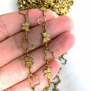 Könnte beinhalten: Nahaufnahme einer goldfarbenen Kette mit Schmetterlings- und blütenförmigen Gliedern. Die Kette wird in einer Hand vor weißem Hintergrund gehalten. Die Kette wird wahrscheinlich für die Schmuckherstellung oder als Verzierung verwendet.