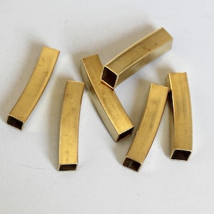 Pode incluir: Seis tubos de metal de cor dourada, alguns curvos, outros retos, com extremidades abertas. Os tubos estão dispostos em um padrão aleatório sobre uma superfície branca.