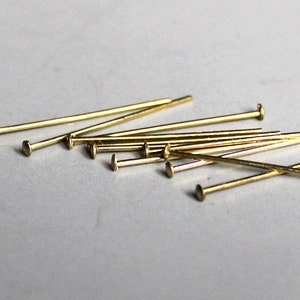 May include: A collection of small, gold-colored head pins scattered on a light gray surface. The pins have a slender, straight shaft and a flat, circular head. These pins are likely used for crafting or sewing projects.