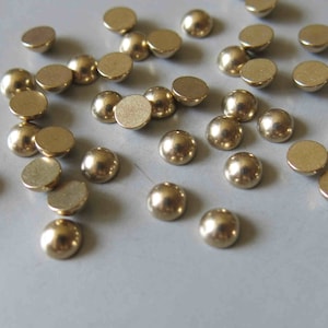 May include: A collection of gold-colored round metal embellishments, some with a domed shape, scattered on a light gray surface.